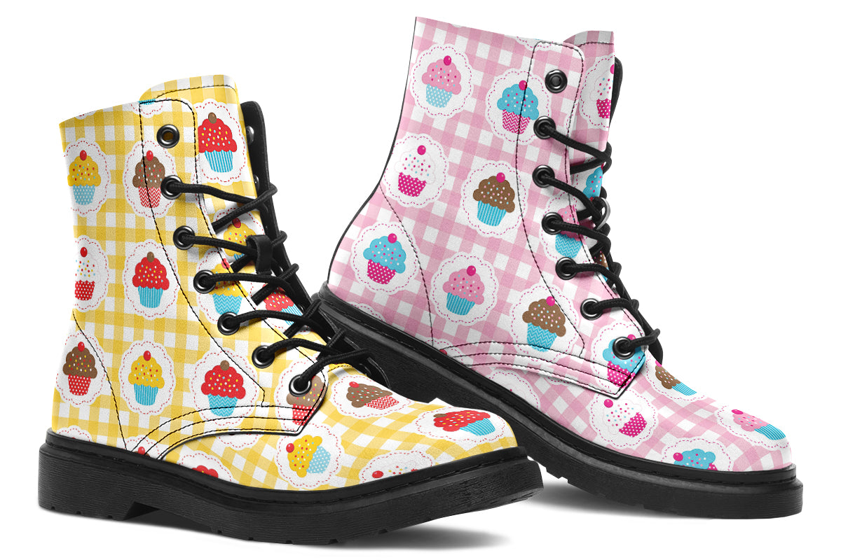 A pair of lace-up combat boots with black soles and laces, featuring a gingham pattern with colorful cupcake illustrations. One boot is yellow and the other is pink, shown against a plain white background.