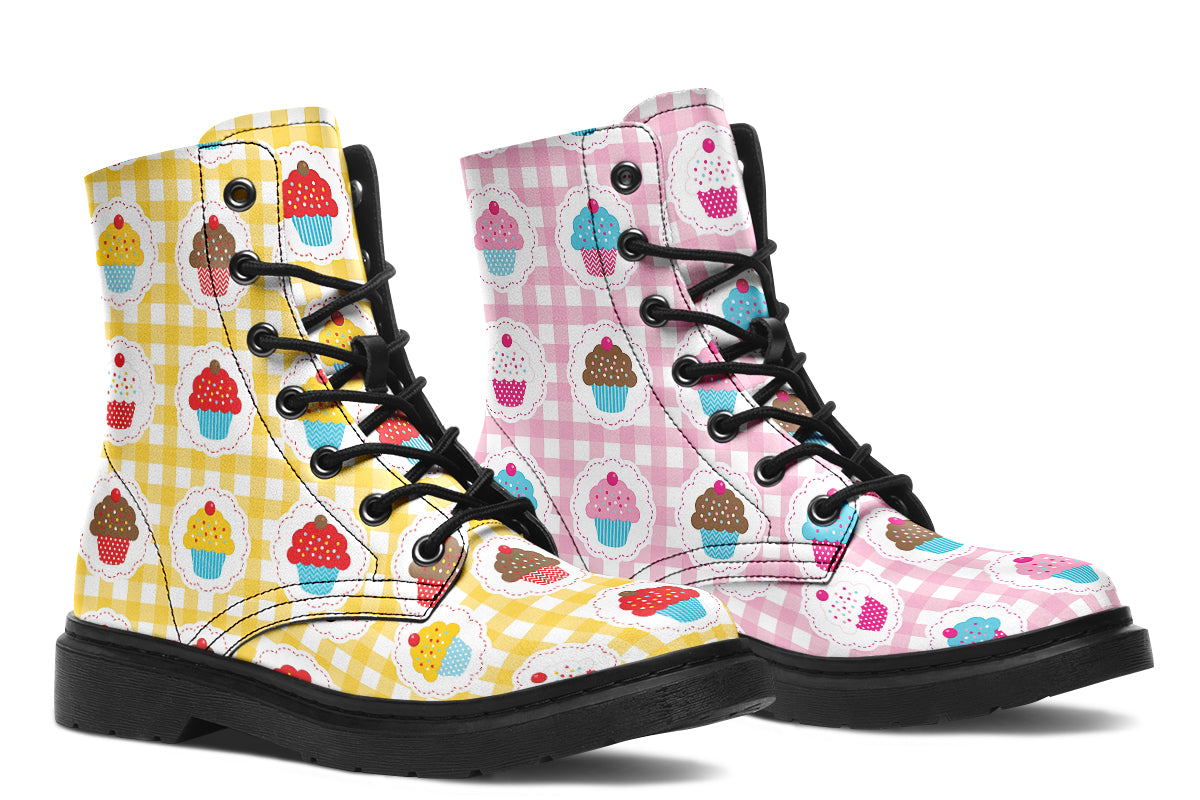 A pair of lace-up combat boots with black soles and black laces, featuring a colorful cupcake print over gingham patterns. One boot has a yellow and white gingham background with multicolored cupcakes, and the other has a pink and white gingham background with multicolored cupcakes.