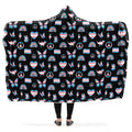 Hooded blanket with a pattern of peace signs, butterflies, dinosaurs, rainbows and heart symbols all in the trans flag colours, on a black background.