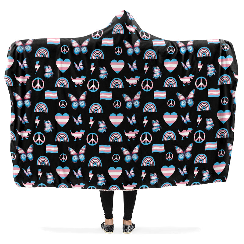 Hooded blanket with a pattern of peace signs, butterflies, dinosaurs, rainbows and heart symbols all in the trans flag colours, on a black background.