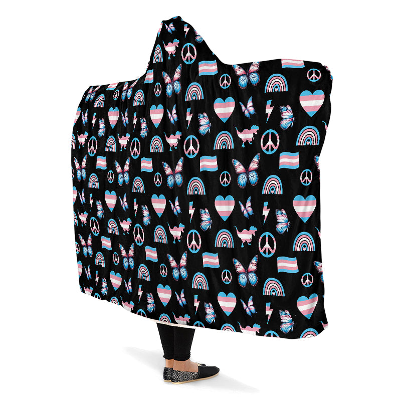Person wearing a hooded blanket with a pattern of peace signs, butterflies, dinosaurs, rainbows and heart symbols all in the trans flag colours, on a black background.