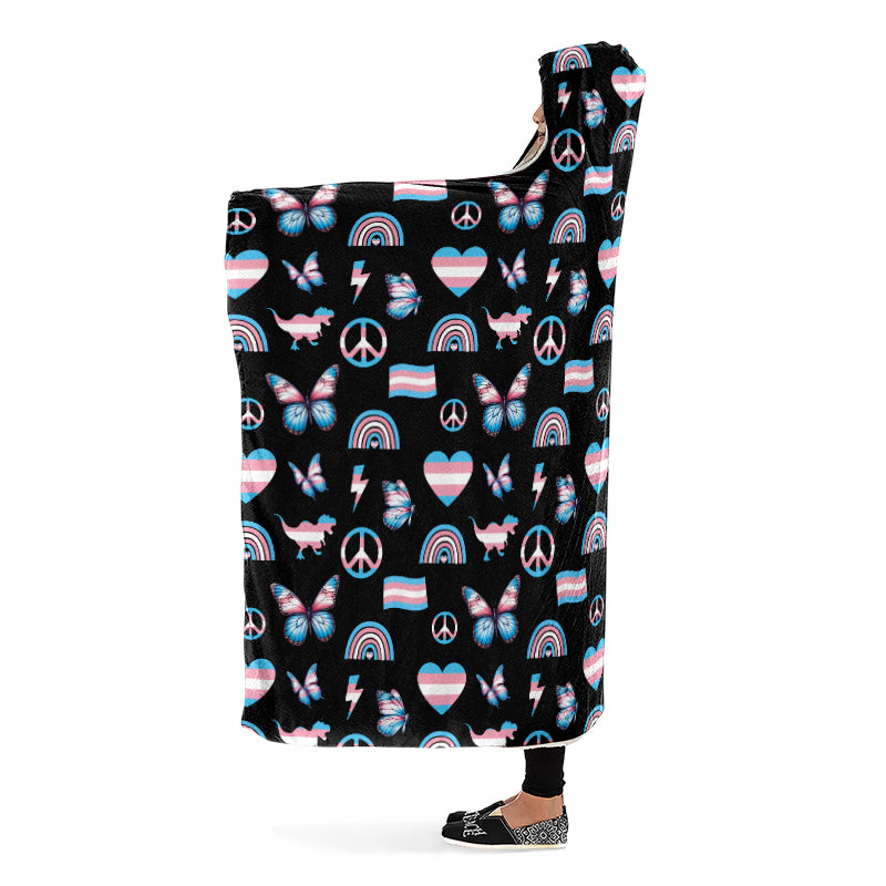 Person wearing a hooded blanket with a pattern of peace signs, butterflies, dinosaurs, rainbows and heart symbols all in the trans flag colours, on a black background. Side view.