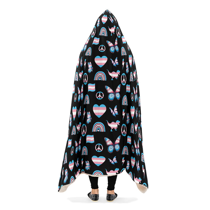 Person wearing a hooded blanket with a pattern of peace signs, butterflies, dinosaurs, rainbows and heart symbols all in the trans flag colours, on a black background. They have their arms down, causing the blanket to drape.